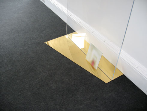 detail of sculptural arrangement gold mirrored perspex on floor with blurred image resting upright against a vertical plane of clear perspex